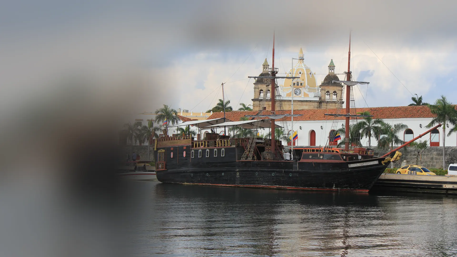 A black pirate-style tour ship with two tall masts moored at a pier in Cartagena, with a yellow church dome and a white colonial building with a red tile roof behind it.