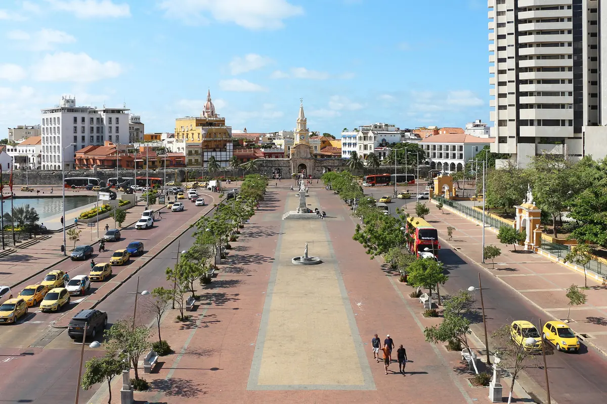 The Clock Gate of Cartagena seen from above at the end of a tree-lined avenue, with yellow taxis on the road.