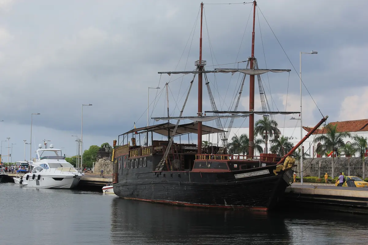 A black two-masted pirate-style tour ship at a pier beside a stone wall, with a yacht and palm trees behind it.
