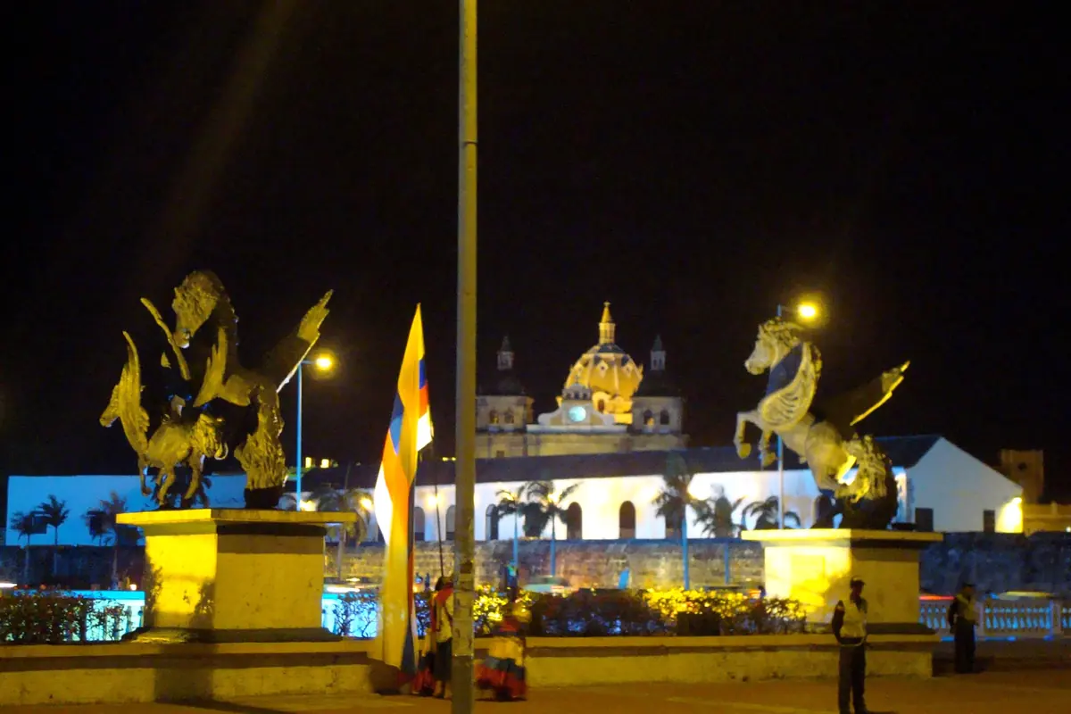Two gilded winged-horse statues on yellow plinths at night, with a church dome lit up behind them.
