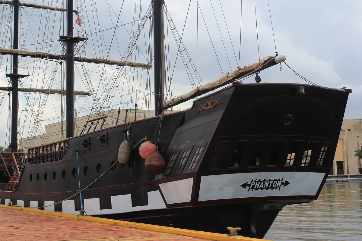 The black bow of a pirate-style tour ship with a white and black chequered band, moored beside a modern building.